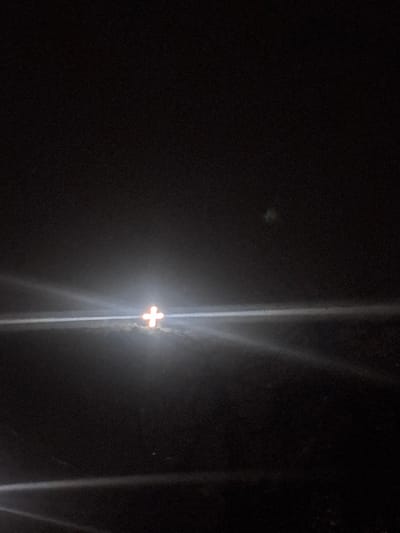 The image captures an outdoor night scene in Lehvaz, Armenia. A brightly illuminated cross-shaped object, emitting a yellowish-orange light, is centered in the mid-ground. Strong horizontal light flares emanate from this source, particularly extending to the right across the frame. The area immediately surrounding the base of the cross is bathed in its light, appearing to rest on a low, indistinct elevation or terrain feature, partially obscured by atmospheric haze or mist. The surrounding environment is predominantly dark, with no other discernible structures or individuals visible. A faint, diffuse greenish light or artifact is present in the upper right quadrant.