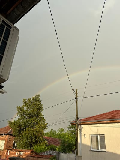 The image depicts a residential area in Morava, Bulgaria, under a sky displaying a faint double rainbow. The view is from a low angle, featuring the tops of buildings and a tree. Power lines intersect the scene. A light-colored building with a window and a red-tiled roof is on the left. A brick building with visible window openings is partially seen at the top left. A white air conditioning unit is visible at the top right of the frame.