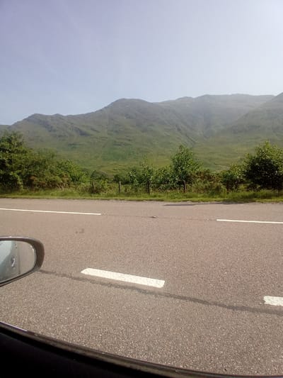 The image shows a view from a car window, looking out at a mountain landscape. The foreground is dominated by an asphalt road with a white line marking the lane. The road appears to be a two-lane highway. To the left, a portion of the car's side mirror and window frame is visible. Beyond the road, there is a strip of green vegetation, including trees and bushes, with a fence line running through it. In the background, a large, green, grass-covered mountain range rises into a pale blue, hazy sky. The overall impression is of a scenic drive through a rural, mountainous area. The location is specified as Kyle, United Kingdom, suggesting a setting in the Scottish Highlands.