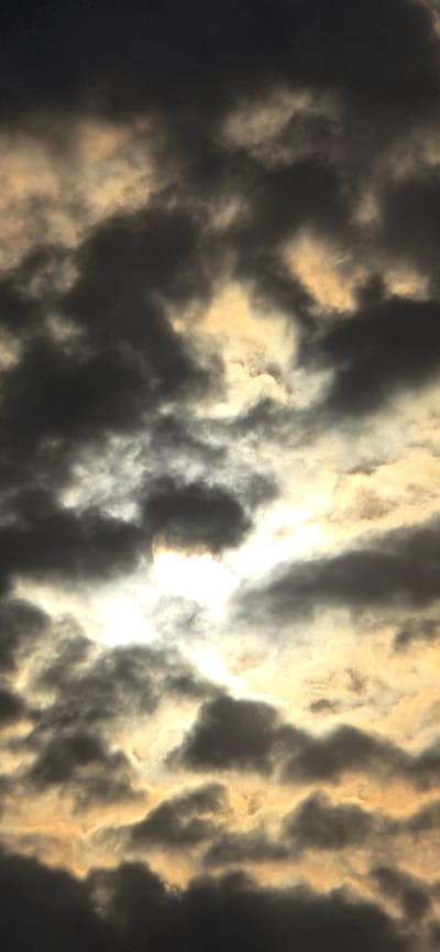 This is a vertical, close-up, low-angle photograph of a cloudy sky. The clouds are dark and ominous in the foreground and at the top, with brighter, golden-yellow clouds interspersed throughout the midground. The sun is visible, peeking through a gap in the clouds, creating a bright, hazy effect. The clouds are varied in texture and density, with some appearing wispy and others more solid. The overall impression is one of dramatic and turbulent weather. The location is in the city of Русская Деревня-2 Кп, Russia.