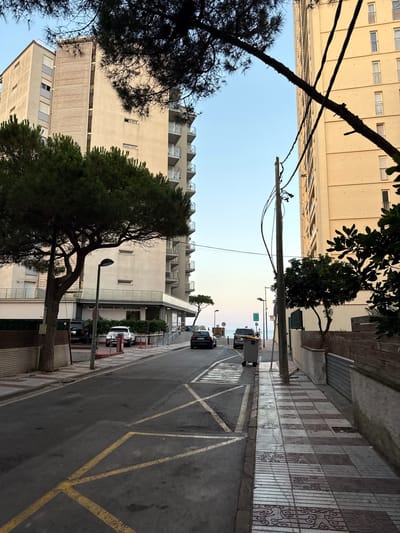 This media file depicts an outdoor scene with a street running parallel to a sidewalk and large buildings. The perspective is from a low angle, looking upwards towards the sky and across the street. The scene appears to be in a residential or tourist area, possibly a coastal town given the distant view of the sea and the style of architecture, which is consistent with Platja D'aro, Spain.

The primary subjects include several multi-story buildings with balconies, some with awnings. Trees, including a large pine tree with dark needles and a smaller leafy tree, are prominent in the foreground and along the sidewalk, casting shadows. The street is paved and marked with yellow lines, suggesting it is a road with parking or traffic lanes. A few cars are visible on the street, including a dark SUV and a white vehicle. A yellow taxi is also present.

The setting is urban, with a tiled sidewalk featuring decorative patterns and a concrete wall. Electrical wires and poles are visible overhead, indicating infrastructure. A lamppost stands on the sidewalk. In the distance, the sky is clear and pale blue, transitioning to a soft orange or pink hue near the horizon, suggesting it is either early morning or late afternoon. The overall atmosphere appears peaceful and quiet.

Notable details include the architectural style of the buildings, the presence of trees, and the markings on the road. No people are clearly visible in the image, and the focus is on the environment and structures. There is no visible text in the image. The time of day is likely dawn or dusk due to the sky's coloration. The weather appears to be clear and pleasant.