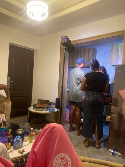 The image shows an indoor scene, likely in Ado-Ekiti, Nigeria, given the prompt. Two individuals are standing; one wearing a dark t-shirt and jeans with decorative stitching on the back pocket, and the other wearing a light green shirt and a blue baseball cap. It appears the first person is attending to the other in some way. There is lighting equipment set up, suggesting this might be a photoshoot or video shoot. A table with various items, including water bottles, snacks, and cosmetics, sits in the foreground. The interior space includes a door and what appears to be another room visible through an opening. A pink garment is draped in the lower right corner.