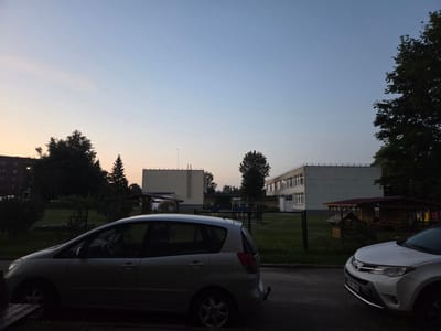 The image shows an exterior view of a street in Širvintos, Lithuania, during dusk. Two cars are parked on the side of a paved road. In the foreground, a silver hatchback is parked on the left. To the right, a white SUV is partially visible. Behind the cars, there is a fenced area containing a playground with some wooden structures. Two multi-story buildings are visible in the background, appearing to be a school or residential complex. The sky is clear and shows a gradient of light blue to pale yellow near the horizon. There are several trees and some vegetation present in the scene. The lighting suggests it is either early morning or late evening.