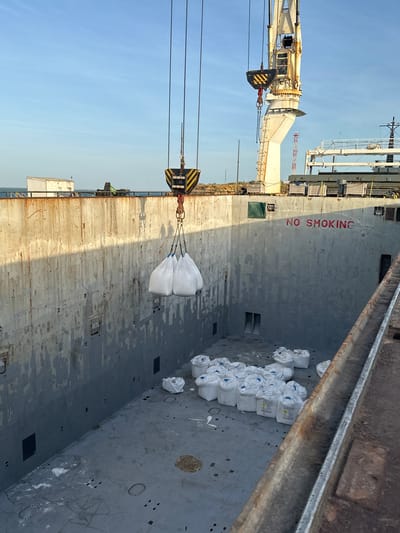 In the city of Uribia, Colombia, a cargo ship's hold is being loaded with large white bags. Three bags are suspended by cables from a crane, about to be lowered into the hold. The hold is empty except for a large pile of similar white bags already on the floor. The interior of the hold is made of grey, weathered metal walls. A red "NO SMOKING" sign is visible on the right wall. The sky is clear and blue.