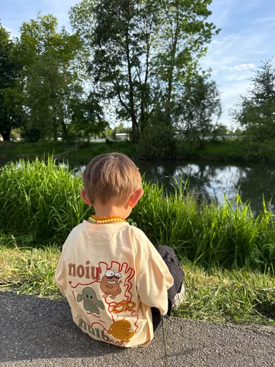 A young person is sitting on a pavement edge in Riedlingen, Germany. Behind them is a river bank with tall grass and a tree line. The person wears a light-colored shirt with cartoon graphics and a beaded necklace.