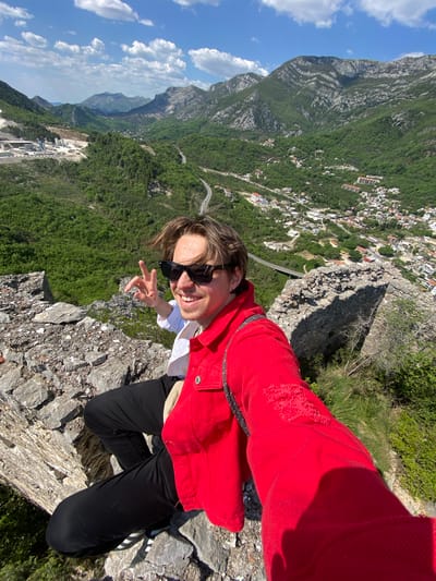 A person is captured in a high-angle selfie, kneeling on a rocky cliff overlooking a vast, mountainous landscape. The individual, appearing male, is wearing a vibrant red jacket with some distressed details on the sleeve, a white shirt, black pants, and sunglasses. They are smiling broadly at the camera and making a peace sign with their right hand, conveying a sense of joy and excitement.

The background reveals a dramatic and scenic vista. Below the cliff, a densely forested valley stretches out, dotted with a settlement featuring numerous white-roofed buildings, indicative of a town or village nestled among the green hills. A winding road can be seen snaking through the landscape. Further in the distance, more rugged mountains rise under a clear blue sky with scattered white clouds, suggesting good weather. Some industrial-looking structures are visible on a flatter area of the terrain in the distance. The scene captures a bright, sunny day, likely during midday, given the clear lighting and strong shadows. The overall setting implies a high-altitude viewpoint, possibly a hiking trail or a scenic overlook in a mountainous region like Zgrade, Montenegro.