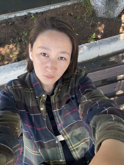 A woman sits on a wooden bench, facing the camera. She has short, dark brown hair and is wearing a green, blue, and red plaid flannel shirt over a black top, and a silver necklace. She has white wireless earbuds in her ears. The bench is outdoors, next to a tree and a planter with mulch. The woman is holding her phone to take the picture, as indicated by the arm extending towards the camera. The environment appears to be a public space, possibly a park or a street with benches. The time of day seems to be daytime, with shadows visible. The background shows dirt, plants, and concrete. The location is stated to be Pereyaslavka, Russia, though this cannot be confirmed from the image alone.