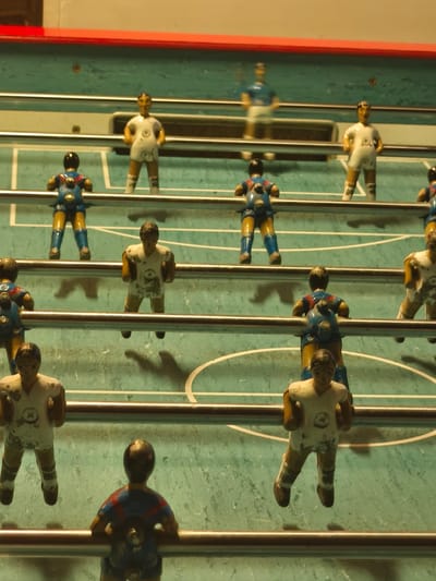 The image shows a well-worn foosball table with painted figurines of soccer players attached to metal rods. The figurines are arranged in rows, with players in white and blue jerseys. The table surface is green with white lines marking the playing field. The background is blurry and out of focus, providing minimal contextual information about location, but given the request for a location and information the image may have been taken in or near Moaña, Spain.