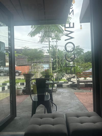 The image presents a view looking outwards through a large glass window or door of an establishment, likely a cafe, in Sukabumi, Indonesia. The perspective is from inside, capturing both interior elements and the external environment.
Inside, on the right side of the frame, are two grey, tufted, upholstered armchairs or stools. Adjacent to the window, a dark metal chair and a table with a glass top are visible. Reflections of the interior ceiling or wall can be seen faintly on the upper left of the glass.
The outside scene, viewed through the glass, features abundant tropical vegetation, including tall palm-like trees and large-leafed plants, indicating a warm climate. The sky appears overcast, suggesting a mild daytime. A concrete and reddish-brown paved area transitions to a street or pathway lined with striped concrete barriers. A dark SUV with a rear-mounted spare tire is parked further down the road, and a building with a tiled roof is visible in the background, nestled among more trees. Power lines are visible against the sky.
Prominently displayed on the glass, visible as reversed text from the inside, is the large word "WELCOME." Other smaller, reversed text fragments are also discernible, including "Coffee," "Espresso," "Latte," "Cappuccino," and phrases such as "We've all been there!", "start my day," and "It's 10 o'clock," likely indicating menu items or cafe slogans. No people are visible, and the scene appears calm.