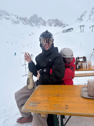 This image depicts an outdoor scene in a snow-covered mountain environment, identified as the city of Klösterle, Austria. Two individuals are seated at a rustic wooden picnic table partially covered in snow. The prominent individual in the foreground, male, wears a black ski jacket, light brown ski pants, and brown ski boots. He is equipped with a black Bern ski helmet featuring a mounted action camera, mirrored Oakley goggles, and a grey neck gait. A white substance is visible on his nose and upper lip. He holds a lit cigarette in his right hand. A beige mitten hangs from his left wrist. The second individual, positioned behind and embracing the first, wears a red ski jacket and a light grey ski helmet with goggles. On the table, to the right, are two empty glass mugs on a small wooden tray. Behind the individuals, another wooden bench or table holds a wooden crate containing multiple dark bottles, one of which appears to be Coca-Cola, alongside several clear plastic cups. Ski poles are visible in the snow to the left of the table. In the background, a vast snow-covered mountain range with jagged peaks is visible under an overcast sky. Skiers are discernible on a snow-covered slope, and a ski lift station with multiple support towers extends into the distance. The wooden table displays a "Stiegl" brand logo.