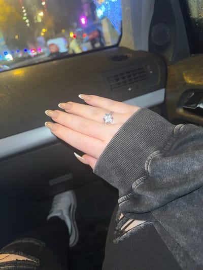 The image depicts a close-up view from within a vehicle at night in Bayraklı, Turkey. A person's left hand, featuring manicured nails with a neutral base and white tips, is positioned in the center-right. A silver-colored ring with a prominent oval-cut clear stone and smaller accent stones is worn on the ring finger. The hand emerges from a dark grey, ribbed long-sleeved garment. In the lower-left, a portion of the person's leg is visible, dressed in dark, distressed jeans with a rip on the thigh, and a white athletic shoe. The dark dashboard of the vehicle is visible across the foreground. Through the windshield, a blurred exterior city scene is apparent, illuminated by numerous scattered, colorful lights (red, blue, yellow, white, magenta), suggesting streetlights, traffic signals, or building lights. Moisture or rain droplets are visible on the windshield surface.