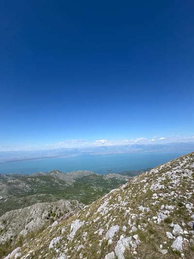 The image shows a mountain landscape in Mali Mikulići, Montenegro, with a view towards the sea. The sky is clear and blue. A rocky mountainside occupies the right side of the frame, with sparse vegetation. In the distance, there is a body of water reflecting the sky, with land faintly visible on the opposite shore.