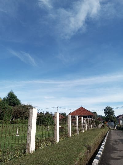 The image depicts an outdoor street scene in Salai Har Htan, Myanmar, under a clear blue sky with scattered wispy white clouds. In the foreground and midground, a fence composed of regularly spaced white concrete posts and rusty dark metal panels extends diagonally from the lower left towards the mid-right. To the left of this fence lies a manicured green lawn area with various trees and shrubs, including a dark green conifer on the far left. To the right of the fence, a narrow, trimmed strip of grass borders a dark asphalt road, which runs towards the background on the right. A black and white striped curb delineates the edge of the road. No individuals or vehicles are visible within the frame. In the background, behind the fence and trees, a building with an orange-red tiled gabled roof is visible. Overhead utility lines traverse the midground horizontally. Further down the road on the right, a white sign with dark lettering, possibly "BPMP," is partially visible. The overall environment appears well-maintained and devoid of active human presence.