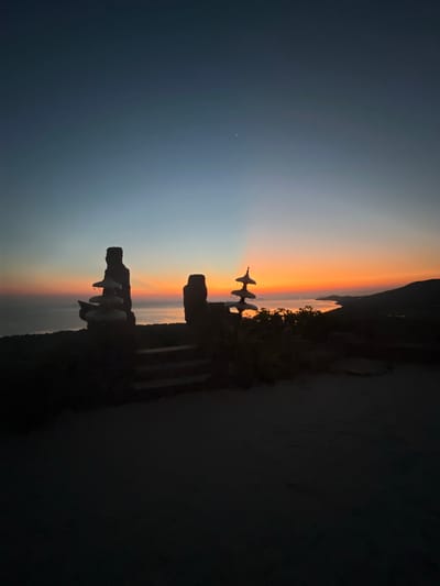 This is a vertical, low-angle, medium shot of a silhouetted landscape at dusk, likely taken from a high vantage point overlooking the ocean. In the foreground, two distinct stacked rock formations are visible, resembling abstract sculptures or cairns. The formation on the left is taller and more slender, with several flat, disc-like stones stacked atop each other. The formation on the right is shorter and wider, with a more irregular shape. Between these formations, a set of dark, stone steps descends into the foreground. The ground in the immediate foreground is dark and appears to be made of dirt or gravel.
The middle ground features a body of water, likely the ocean, reflecting the vibrant colors of the sunset. A distant coastline with rolling hills is visible on the right side of the frame. The sky transitions from a deep indigo blue at the top to a bright orange and yellow band just above the horizon, with subtle rays of light emanating from behind the distant hills. A single, faint star is visible in the upper left portion of the sky. Dense foliage is silhouetted against the bright horizon in the middle ground, surrounding the rock formations.
The scene appears to be located in the Karimunjawa Islands of Indonesia, a popular destination known for its natural beauty and coastal landscapes. The atmosphere is serene and tranquil, characteristic of a sunset in a tropical or coastal region. There are no people or animals visible in the image. The overall impression is one of natural beauty and a sense of peaceful solitude.