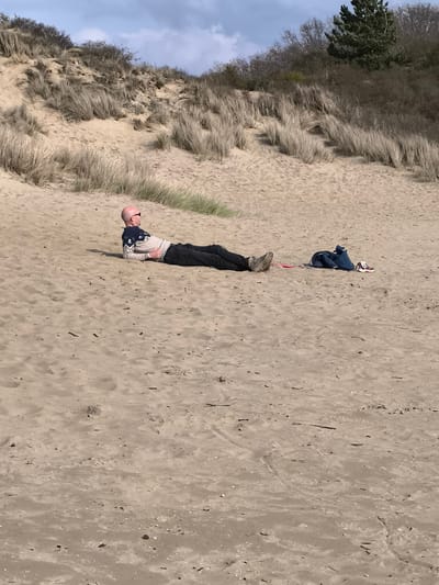 A man lies prone on a sandy beach near Overveen, Netherlands. He wears a patterned sweater, dark trousers, and shoes, and sunglasses. A denim jacket, a pair of sneakers, and a red disc lie beside him. The background consists of sandy dunes covered with dune grass and some trees. The sky is visible.