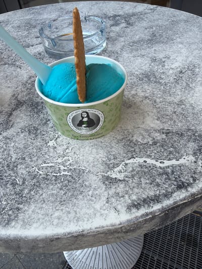 The image shows a close-up view of a table with a dessert and an ashtray. The table has a marbled gray and white surface. In the foreground, there is a white paper cup filled with two scoops of bright blue ice cream. A long, thin, textured cookie is inserted vertically into the ice cream. A pale blue plastic spoon rests on the rim of the cup, its handle extending out of the frame. Behind the ice cream cup, there is a clear glass ashtray with some residue. The background shows more of the marbled tabletop and a hint of the base of the table. The overall impression is a casual outdoor setting, possibly a cafe or restaurant, in Lörrach, Germany. The ice cream cup has a design that includes a circular emblem with a portrait of the Mona Lisa and the text "Mona Lisa" and "Ihre Eismanufaktur für Passion & Genuss".