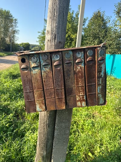 In the village of Bolshiye Shady, Russia, a weathered, multi-unit mailbox system, showing significant rust and peeling blue paint, is attached to a wooden post. The mailboxes are arranged in a row, each with a small opening for mail and a lock. The surrounding environment is rural, with lush green grass, trees, and a dirt road visible in the background. A bright turquoise fence can be seen to the right. The sky is clear and blue, indicating a sunny day. No people are present in the image.