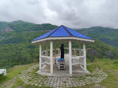 A medium shot depicts a white gazebo with a blue tiled roof situated on a cobblestone area near a green hillside. Inside the gazebo, a person in a dark hooded garment stands on a wooden pallet with a blue plastic bag and red container on it. The gazebo has white pillars and railings. The background features a landscape with green hills and mountains under an overcast sky. The location is identified as Bov, Bulgaria, based on the image context.