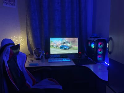 This media file depicts a gaming setup in a room with blue ambient lighting. The scene is set in a dimly lit room, likely at night, with predominantly blue and purple tones.
The main subjects include a gaming chair in the foreground, predominantly black with red and white accents. To the right of the chair is a desk with a computer monitor displaying a blue car driving through a road lined with cherry blossom trees. A white mechanical keyboard with blue backlighting sits in front of the monitor. To the right of the keyboard are two red speakers and a black mousepad with a mouse. A gaming PC tower, lit with colorful RGB fans, is positioned on the desk to the right of the monitor. Beside the PC, a round mirror or frame is visible on the wall. On the left side of the desk, there's a water bottle, headphones on a stand, and a small blue object. A delicate string of warm white LED lights, shaped like a tree, is placed on the desk to the left, illuminating the area around it. A blue curtain hangs behind the desk and monitor, contributing to the overall mood. A framed artwork with a traditional pattern is visible on the upper left wall.
No specific activity is clearly depicted beyond the setup itself, suggesting a space prepared for gaming or work. The overall atmosphere is calm and focused, with the blue lighting creating a modern and immersive environment. The location cues are minimal, but the context provided states the location is Ardon, Russia. There is no visible text in the image.