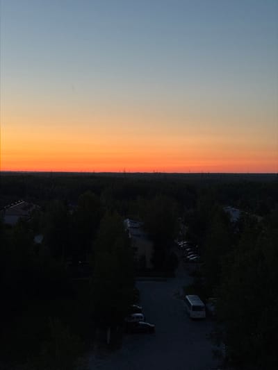 The image depicts a twilight scene over a cityscape, likely Pyt'-Yakh, Russia, given the context. The sky exhibits a gradient of colors, transitioning from a pale blue at the top to a vibrant orange near the horizon, indicating either sunrise or sunset. Silhouetted against the horizon are numerous tall, thin structures, possibly power line pylons or radio towers, stretching across the landscape. Below the sky, a dense canopy of dark trees occupies the midground, obscuring much of the urban environment. Within the treeline, glimpses of buildings and rooftops are visible, suggesting a suburban or residential area. In the foreground, a street or courtyard is dimly lit, with several vehicles parked along the side. One vehicle, a white bus, is prominently positioned in the lower right quadrant. The overall atmosphere is one of fading light and quiet urban presence.