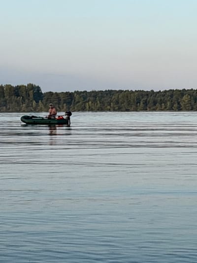 In the city of Gurino, Russia, a man is seen on a small, inflatable boat with an outboard motor on a calm body of water. He is wearing a light-colored cap and appears to be shirtless. The boat is dark green and has an orange life vest inside. The water is rippled, suggesting a gentle breeze. In the background, a dense line of trees forms the horizon, with a light blue sky above. The overall scene depicts a tranquil moment on the water.