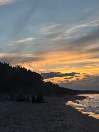 The image depicts a beach scene at dusk, likely in Plieņciems, Latvia, with a vibrant sunset painting the sky. The sky is filled with wispy clouds, transitioning from a clear blue on the left to warm hues of orange and yellow closer to the horizon, indicating the sun is setting. A line of dark trees silhouettes the land against the colorful sky.

In the foreground, a sandy beach stretches out. Several people are gathered on the beach, some seated in chairs, facing towards the water and the sunset. They appear to be enjoying the view and the end of the day. There's a pile of wood that might suggest a bonfire, although no fire is visible. The water's edge is visible on the right, with gentle waves reflecting the sunset's light. The overall mood is peaceful and serene, capturing a moment of relaxation and appreciation for nature. The time of day is clearly evening, as evidenced by the sunset and the fading light casting shadows. No text is visible in the image.