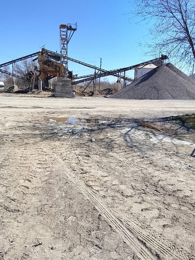 The image depicts an aggregate processing facility located in Yangi Chinoz, Uzbekistan, under a clear blue sky. The scene is observed from ground level, looking towards the operational machinery.
In the immediate foreground, the ground consists of dry, light-brown soil mixed with darker, damp patches, suggesting recent moisture. Numerous tire tracks, predominantly from heavy vehicles, are distinctly visible traversing the soil, along with scattered boot prints. A notable muddy puddle reflects the blue sky near the center-left. A horizontal section of rusty metal mesh or rebar lies on the ground in the mid-right foreground.
The midground is dominated by industrial infrastructure. A large, complex arrangement of conveyor belts, supported by steel trusses and concrete pillars, forms the core of the aggregate processing plant. These belts converge on and emanate from a central, multi-level yellow and grey metal structure, likely housing crushers or screening equipment. A tall, narrow steel tower with a platform is visible on the central structure. To the right of the machinery, a substantial, dark grey conical pile of gravel or crushed aggregate stands, partially obscuring a two-story building with a light-colored facade and a red-tiled roof in the background.
To the left of the main processing equipment, a white vehicle (possibly a van or truck cab) is partially visible. Sparse, bare tree branches extend into the upper right corner of the frame, casting shadows on the ground in the mid-right. Power lines are visible stretching across the upper midground. No individuals are present, nor are there any signs of active operations. The overall impression is that of an industrial site in a state of disuse or temporary inactivity on a bright day.
