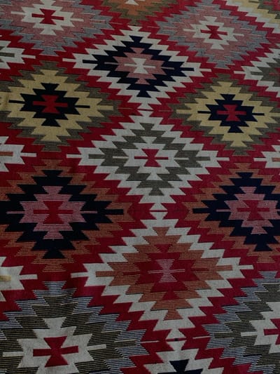 The image displays a close-up, overhead view of a multi-colored woven textile, identified as being in Yasinia, Ukraine. The fabric, likely a rug or tapestry, features a repeating geometric pattern composed of interlocking diamond and zigzag motifs. Dominant colors include shades of red, light beige, dark blue, and various tones of brown and gold. The textile exhibits a visible woven texture with horizontal striations evident in some color blocks. No individuals, specific actions, or other objects are present in the scene, which appears to be a flat, uniformly lit surface.