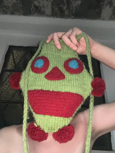 A person is holding a knitted fish-shaped bag. The bag is primarily green with red and blue accents. The fish features round blue eyes outlined in red, a red triangle, and a red stripe down its side. Three red pom-poms act as fins. The bag has a green knitted strap for carrying. The background shows a window and a wall, but the focus is clearly on the knitted bag.