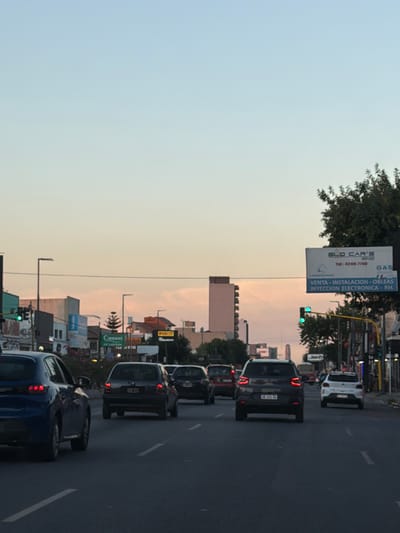 A street scene at dusk features congested traffic. Cars line the right side of the frame, moving in the same direction. In the background, buildings and structures are silhouetted against the fading sky. Street signs, including "Cannod" and "ELO CAR'S," are visible on the left. A traffic light shows a green signal. The sky transitions from light blue to a soft orange hue towards the horizon.