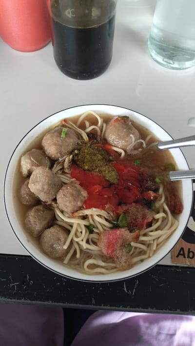 A close-up overhead shot captures a bowl of Indonesian noodle soup, likely Bakso, positioned on a white table. The dish features a generous portion of broth, thick yellow noodles, and numerous meatballs. Visible toppings include a dollop of vibrant green chili paste (sambal) and a smear of red chili sauce. The bowl itself is white with a black rim. To the left of the bowl, a red bottle is partially visible, and next to it, a dark glass bottle containing a dark liquid, possibly soy sauce or chili oil, stands prominently. A glass of clear water is situated to the right of the dark bottle. The scene is set in what appears to be a restaurant or home dining area in Panteraya, Indonesia. The table surface is white and appears clean, with subtle shadows cast by the objects. In the bottom right corner, a wooden sign with the partial word "Ab" is visible. The lower portion of the image shows the upper thighs of a person wearing purple clothing.