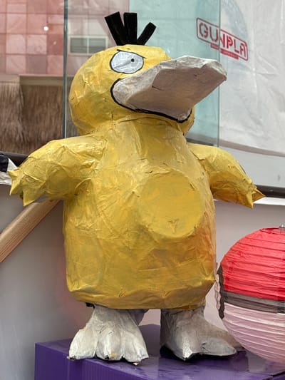 A yellow, paper-mâché figure of the Pokémon Psyduck stands on a purple rectangular platform in El Paso, United States of America. The figure is approximately three feet tall and appears to be a piñata, evidenced by the textured paper covering and the visible seams. Psyduck's beak is made of a light gray, crinkled material, and its eyes are large and white with black pupils. The figure has black, spiky hair on its head. To the right of the Psyduck figure is a red and white spherical object, possibly a lantern or another decorative item. The background consists of a glass display window, through which hints of interior elements like shelves and possibly a retail environment are visible. A red and white sign with the word "GUNPLA" in red lettering is partially visible in the upper right portion of the image, suggesting the location may be a store selling models or toys. The lighting is bright, creating reflections on the glass.