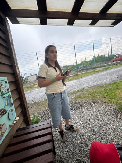 The image depicts a young woman standing under a wooden and translucent roof structure, likely an outdoor shelter or pavilion. She is wearing a light-colored polo shirt with a dark collar and trim, light blue denim shorts, and athletic shoes. The woman is holding a smartphone in her right hand and appears to be speaking, looking slightly to her right. A green wristband is visible on her left arm.
The background reveals a fenced area with a gravel pathway and patches of grass. Beyond the fence, trees and a small building are visible. A red vehicle is parked in the distance. The sky is overcast, suggesting a cloudy day. To the left of the woman, a wooden wall with a colorful interactive display featuring drawings of birds and gears is attached to the shelter's structure. A dark wooden bench is situated in front of this display. The overall scene suggests an outdoor recreational or educational setting. The location is identified as Zaliznychne, Ukraine.