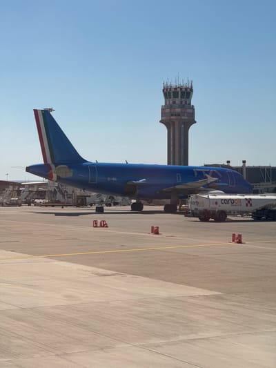 The image depicts an airport scene, likely at Palermo Airport (PMO) in Cinisi, Italy, based on the presence of an ITA Airways aircraft. The aircraft is parked on the tarmac next to a Carboil fuel truck. An airport control tower is visible in the background. Ground markings and safety cones are present on the tarmac. The sky is clear.