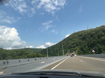 The image captures a view from a moving vehicle on a highway in Kveda Tsiplavake, Georgia. The highway is multi-lane and asphalt, with white lane markings. On the left side of the highway, a concrete barrier with a grey metal railing runs parallel to the road, separating it from a forested hillside. The hillside is densely covered with green trees, characteristic of the region's natural landscape. On the right side of the highway, another forested hillside is visible, featuring two tunnel entrances. One car and a white van are driving on the highway, approaching the tunnels. The sky above is blue with scattered white clouds. Power lines are visible on the higher parts of the hills. A triangular traffic sign with a tunnel symbol is posted on the right side of the road, indicating the presence of tunnels ahead. The overall scene depicts a segment of modern infrastructure integrated into a natural, hilly environment.