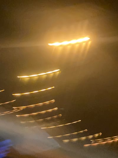 The image depicts blurred streaks of light against a dark, hazy background, suggesting a long exposure photograph of a nighttime scene. The dominant colors are warm yellows and oranges from the lights, contrasted with a dark brown or sepia tone of the environment. Several horizontal and diagonal streaks of light are visible, some appearing as broken lines and others as continuous bands. One prominent band of light is located in the upper portion of the image, fanning out into smaller streaks. Below this, a series of parallel, slightly curved light streaks descend diagonally towards the bottom right. A soft, misty or smoky element is present in the lower left, with faint hints of blue. The overall impression is abstract and impressionistic, lacking clear identifiable objects or people. It is not possible to determine the specific location within the city of Chesapeake, United States of America, from this image alone.