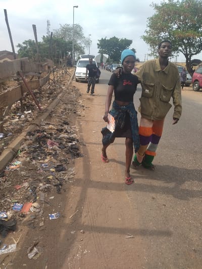 The image shows two young adults, a man and a woman, walking together along a paved road in the city of Maraba, Nigeria. The woman is wearing a blue beanie, a black t-shirt with the word "Barbie" printed on it, a black and blue skirt and pink sandals. The man is wearing a green jacket, a white t-shirt, a multicoloured knitted skirt and black sandals. The man has his arm around the woman's shoulders. The road is lined with a wooden fence on one side and a large pile of garbage on the other. The garbage is composed of mostly plastic and paper. The road is relatively empty with some other people visible in the background. The sky is mostly cloudy.  The coordinates of the location are 9.024 latitude and 7.573 longitude.