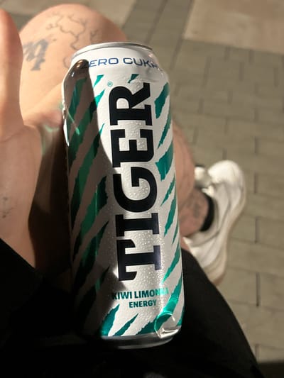 A young adult, likely male given the tattoos and clothing, is holding a can of "Tiger" energy drink. The can is silver with a green and white striped design, and the word "TIGER" is prominently displayed in black lettering. Text on the can indicates "ZERO CUKRU" and "KIWI LIMONKA ENERGY". The person is wearing black clothing and white sneakers. Tattoos are visible on the person's left arm and thigh, one appearing to be a map outline. The background consists of paved ground, possibly a sidewalk or plaza, with a brick-like texture. The image appears to have been taken outdoors in the evening or at night, given the lighting. The person's left thumb is visible in the foreground, positioned as if to support the can. The overall setting is urban, and based on the presence of the "ZERO CUKRU" text, which is Polish for "zero sugar", the location is likely Poland, specifically the city of Siedlce.