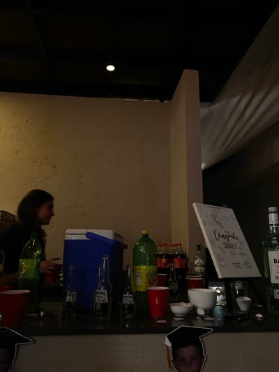 The image depicts a bar scene with various alcoholic beverages and accessories. A "Congrats Charly" sign leans against liquor bottles and ceramic cups. Other objects on the bar include soda, Corona bottles, and a blue cooler. A cardboard cutout of a person wearing a graduation cap is present. The location appears to be an indoor setting, possibly in Santa Cruz Tehuixpango, Mexico. A person is partially visible in the lower-left quadrant of the image.