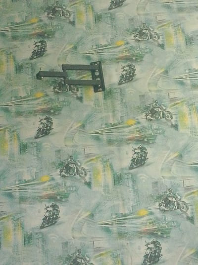 The image displays a light-blue and green patterned wallpaper with a repeating motif of motorcycles in motion. The pattern appears to be a stylized representation of cityscapes with streaks of light, suggesting speed. There are no people, animals, or recognizable landmarks that would indicate a specific location in Asino, Russia. In the center of the image, a dark, multi-jointed bracket is mounted on the wallpaper. This bracket is the only three-dimensional object clearly visible, contrasting with the flat, printed pattern. The overall impression is of a decorative wall covering with a dynamic, urban theme.