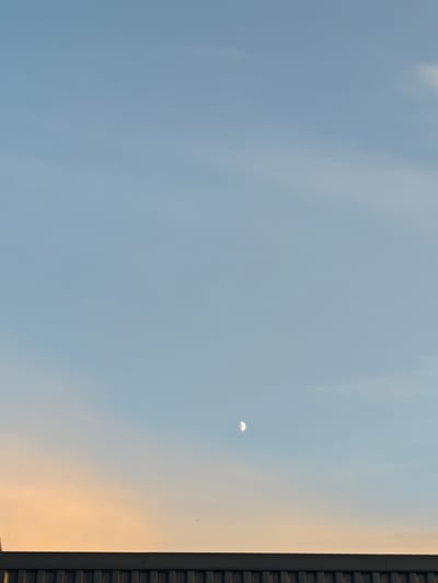 The image shows a clear sky with a waning crescent moon visible. The lower portion of the sky exhibits an orange hue, suggesting it is either sunrise or sunset. A dark, flat structure, possibly a building or a fence, runs horizontally across the bottom of the frame. No people are visible in the image. The scene is set against the backdrop of the city of Kostomarovo, Russia.