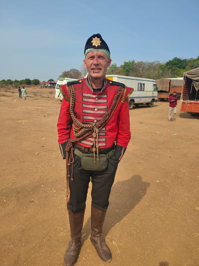 A man in an elaborate red and black military-style costume stands on a dirt field in Vamasamudram, India. The costume includes a red jacket with silver trim, black pants with a red stripe down the side, and brown leather boots. He wears a black hat with a gold emblem on the front. The man has his hands in his pockets. In the background, there are several vehicles, including vans and trucks, and a few people walking around. The sky is clear and blue.