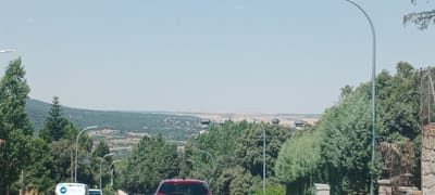 The image captures a roadside view of a rolling landscape under a clear sky, likely on a sunny day. In the foreground, a street with several cars is visible, including a red car in the center and the back of a white van with an "ADT" logo on the left. Trees line the road, with taller, darker green trees on the left and lighter green, more conical trees to the right. Several lampposts with modern, curved designs are also present along the roadside. The middle ground reveals a densely wooded area with a distant view of hills and a more sparsely vegetated, yellowish plain. The horizon shows a hazy, expansive vista. The overall scene suggests a location on the outskirts of a town or city, possibly in a hilly or mountainous region. Given the context provided, this scene is located in El Espinar, Spain.