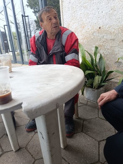 The image presents an outdoor setting in Vinitsa, Bulgaria. An elderly male with short brown hair is seated, oriented right, his mouth slightly open, indicating speech. He wears a red long-sleeved shirt and a red, grey, and black work jacket with reflective banding. Dark work boots with red soles are visible on his feet, resting on hexagonal grey paver tiles. In the foreground, a weathered white plastic round table occupies the central and left portions of the frame. Two plastic cups are on the table, with one in the extreme foreground containing a dark liquid. A second individual's hand and dark clothing are partially visible on the lower right, positioned low to the ground, suggesting a squatting or kneeling posture. The background on the left features large glass panels reflecting trees and an overcast sky. On the right, a textured, light-colored wall with visible wear and a small dark hole serves as a backdrop, with a potted green plant in a rectangular container situated against it.