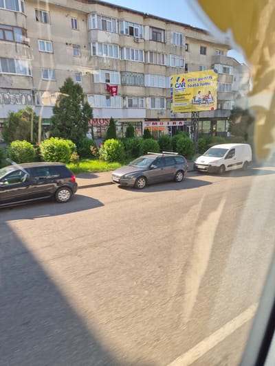 This is a daytime, eye-level, slightly angled outdoor photograph taken from a moving vehicle, looking towards a building in Fălticeni, Romania. The building is a multi-story apartment block with a neutral, light-colored facade, featuring numerous windows and balconies. A large yellow billboard is prominently displayed on the side of the building, advertising "CAR" with the slogan "este pentru toată lumea" (is for everyone) and text in Romanian about financial services. In front of the building, there is a row of green bushes and trees. A paved road runs in front of the building, with three vehicles parked or moving on it. From left to right, a black hatchback car, a dark gray station wagon, and a white panel van are visible. The road and sidewalk show some wear and tear. The image is partially obscured by a yellowish, blurred reflection in the foreground, likely from the vehicle's window.