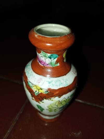 A small, decorative ceramic vase, approximately 10-15 cm in height, is depicted against a dark, indistinct background, resting on a reddish-brown tiled surface in Longmiang, Laos. The vase is flask-shaped with a narrow, slightly flared neck, a rounded body, and a small base. Its surface features a white background adorned with two prominent, horizontal reddish-brown bands. Between these bands and on the upper neck, floral motifs in pink, yellow, green, and light blue are painted. The rim and base are also reddish-brown. A distinct crack is visible on the reddish-brown rim of the vase, extending downwards into the neck. The lighting indicates a localized light source, leaving most of the environment in shadow. No individuals or other objects are present in the immediate vicinity.