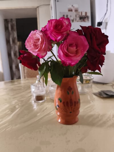 A vase of roses sits on a table. The vase is orange and has green and red floral decorations. The roses are a mix of pink and dark red. In the background, there is a refrigerator with photos on it, and a doorway. The table appears to be in a dining room.