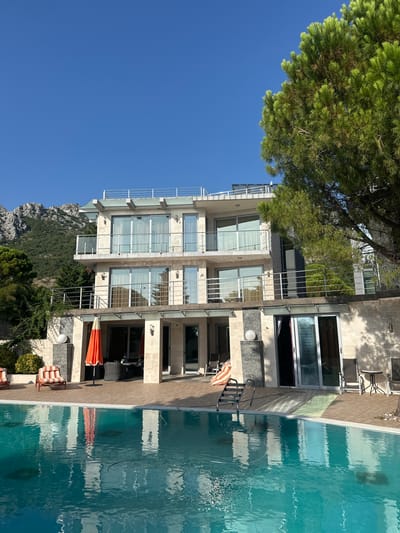 A luxurious, multi-story modern villa with large windows and balconies is situated in a scenic mountain setting. A clear blue sky serves as the backdrop. In the foreground, a swimming pool reflects the building and surrounding greenery. Several lounge chairs are placed near the pool, and an orange umbrella is open. The villa's architecture features a light-colored stone facade and a white railing on the balconies. Lush green trees, including a large pine tree on the right, surround the property, and a rugged mountain range is visible in the distance. The location is identified as Brca, Montenegro.