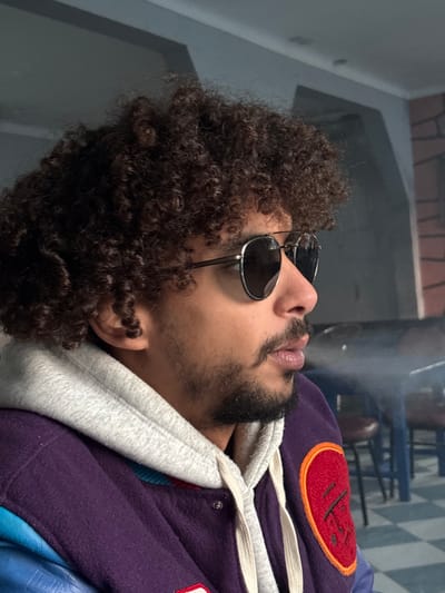 A man with dark curly hair and sunglasses lies on his side. He has a beard and light skin. Smoke or vapor is exhaling from his mouth. He's wearing a purple jacket with a white hoodie underneath. A red and orange patch is visible on the jacket. The background shows a wall and what appears to be furniture indoors.