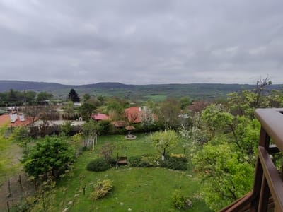 An eye-level, slightly aerial shot shows the backyard of a property in General Kantardjievo, Bulgaria, with a verdant lawn surrounded by trees, shrubs, and several buildings, under a cloudy sky. A wooden railing occupies the right side of the frame, slightly obscuring the view.
In the backyard, a grassy lawn dominates the scene. A seating area with a wooden bench is located in the center of the lawn, bordered by shrubbery. A covered well is visible. A soccer goal net stands on the lawn.
Red-roofed buildings are scattered in the background, interspersed with trees. Beyond the properties, forested hills extend horizontally across the horizon. The sky is overcast with dense clouds.
